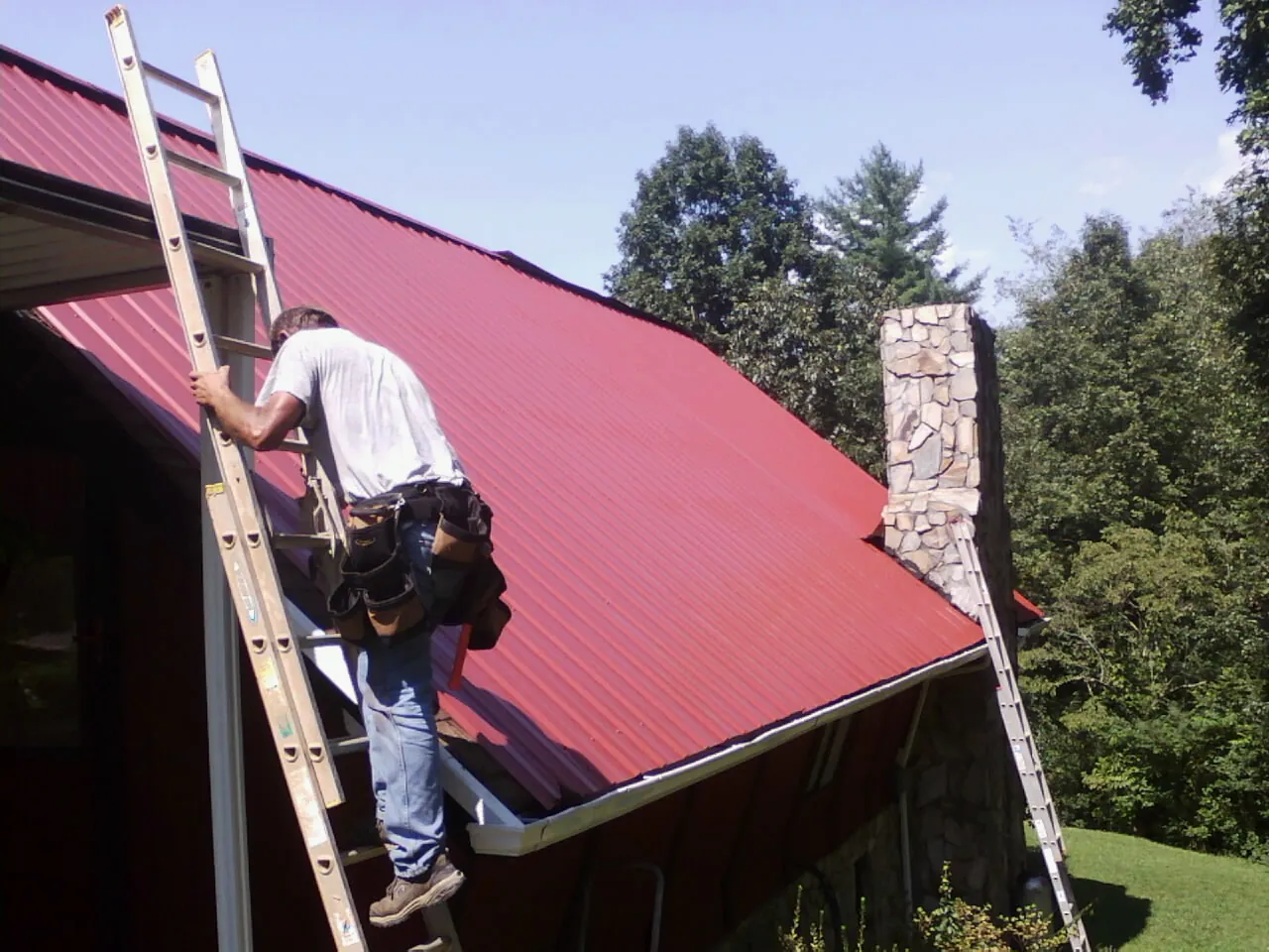 Roofer on a ladder beside a red metal roof and stone chimney — Soffit & Fascia Repair by Roofing Brothers in Rocky River